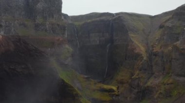 Yeşil bitki örtüsüyle kaplanmış yüksek dik kenarlı derin kayalık kanyon. Geriye doğru uzun şelalenin görüntüsü. Güzel doğal sahne. Haifoss, İzlanda