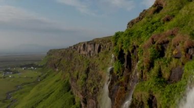 Şelaleli yüksek uçurumlar boyunca gergin bir uçuş. Yeşil bitki örtüsü kayanın üzerinde büyüyor. Seljalandsfoss şelalesinin manzarası güzelmiş. İzlanda