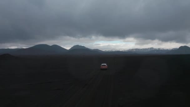 Volar sobre SUV coche de conducción en un paisaje estéril cubierto de cenizas volcánicas. Vista panorámica de las montañas en el fondo y nubes oscuras en el cielo. Islandia — Vídeos de Stock