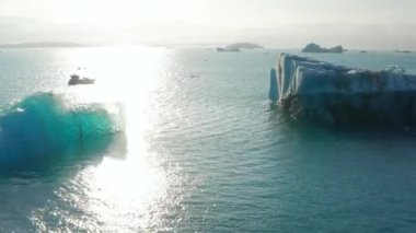 Su yüzeyinde yüzen buz blokları. Geriye doğru buzul parçalarıyla birlikte göl görünüyor. Alçak güneşe karşı görüş. İzlanda