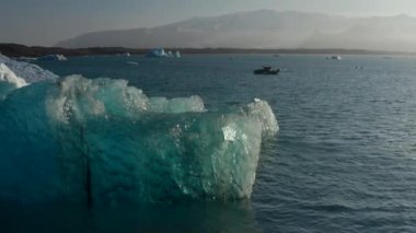 Durgun su yüzeyinde yüzen mavi buzdağı batan güneş tarafından aydınlatılıyor. Arka planda dağ sırtı. İzlanda