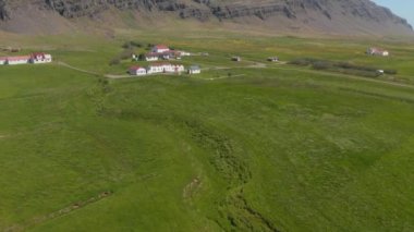 Yeşil çayırın ortasındaki bir grup evin görüntülerini yolluyorum. Arka planda yükselen Rocky dik yamacı. İzlanda