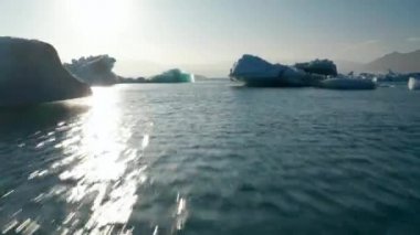 Yüzen buz blokları arasında su yüzeyinden alçak uçuş. Eriyen buzulun parçaları, güneşe karşı görüş. İzlanda