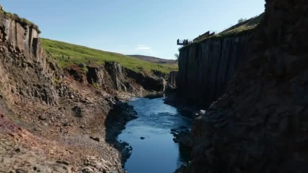 Forwards fly above stream surrounded by rocky banks with basalt columns. Visitors admiring natural attraction. Studlagil Canyon, Iceland — Stock Video