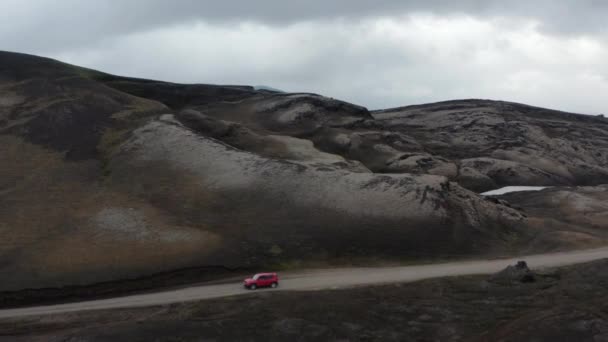 Красная машина едет по грунтовой дороге вдоль вулканического хребта. Открывая большое озеро и горы позади. Исландия — стоковое видео