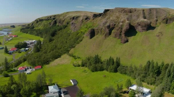Slide και τηγάνι πλάνα από ψηλό γκρεμό με καταρράκτη πάνω από το δάσος και τα κτίρια κατά μήκος του δρόμου. Αεροφωτογραφία τοπίου. Ισλανδία — Αρχείο Βίντεο