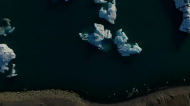 Göl üzerinde yüzen buz bloklarını yukarıdan gören uçan kuşlar. Aydınlanmış yüzeyler, karanlık ve sakin sularla çelişiyor. İzlanda