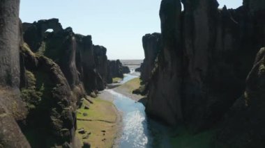 İleriye doğru sakin bir nehirle kanyon boyunca uçar. Güneş ışığına karşı görüş. İnsanlar yamaçta dikiliyorlar. Fjadrargljufur Kanyonu, İzlanda
