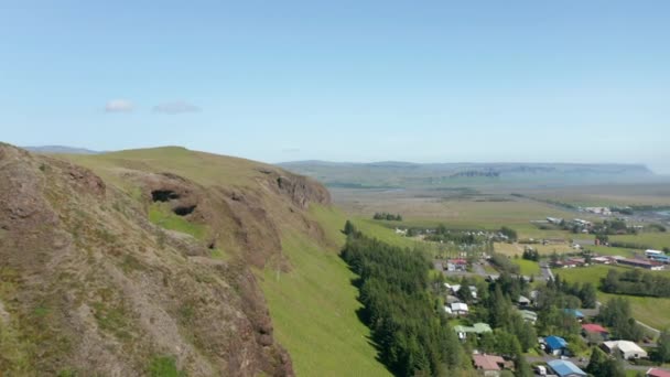 Budynki i przyczepy kempingowe pod osłoną skalną. Naprzód opadająca mucha wzdłuż krawędzi chropowatego zbocza. Islandia — Wideo stockowe