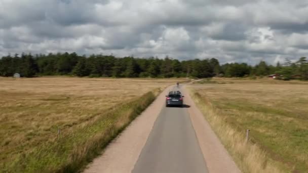 Flyga runt bil kör på smal väg på landsbygden. Cyklisten rider framför bilen. Byggnader i utkanten av skogen under dramatiska moln på himlen. Danmark — Stockvideo