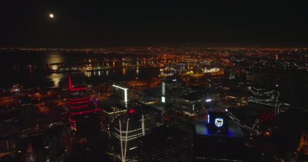 海岸沿いの近代的な高層ビルや港がライトアップされています。後ろの夜の大都市の上を飛ぶ。水面に月の光が反射する。南アフリカ共和国ケープタウン — ストック動画