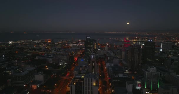 Voorwaarts vliegen boven een rij hoge gebouwen in het centrum van de stad. Zee baai met grote zeehaven aan de kust 's nachts. Kaapstad, Zuid-Afrika — Stockvideo
