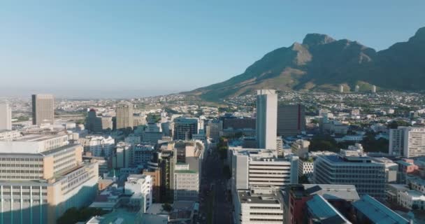 İleri, şehir merkezindeki çağdaş ofis binalarının üzerinden uçuyor. Arka plandaki dağların yamacında yerleşim yerleri yükseliyor. Cape Town, Güney Afrika — Stok video