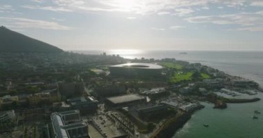 Deniz kıyısının havadan görünüşü ve rıhtımdaki binalar. Günbatımına karşı futbol stadyumunun modern dairesel yapısı. Cape Town, Güney Afrika
