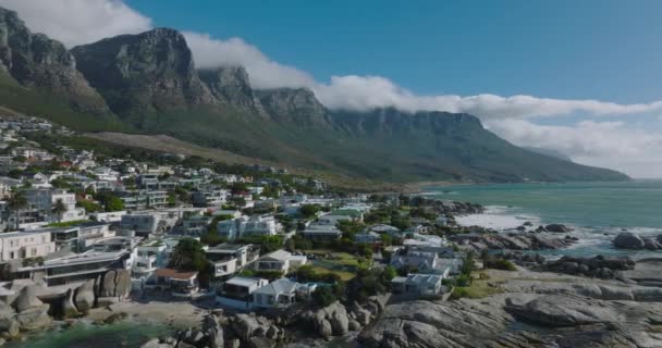 Légi felvételek a tengerparti ingatlanokról. Hullámok mossák a part menti köveket. A fenséges hegygerincet felhők borítják a háttérben. Fokváros, Dél-Afrika — Stock videók