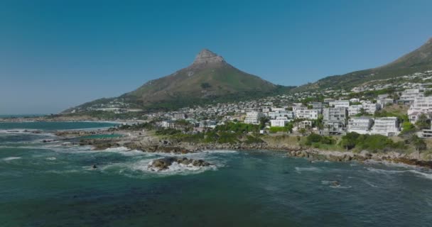 Forwards fly above waves washing sea coast. Residential buildings on waterfront. Tall mountain with steep slopes and rock peak. Clear blue sky. Cape Town, South Africa — Stock Video