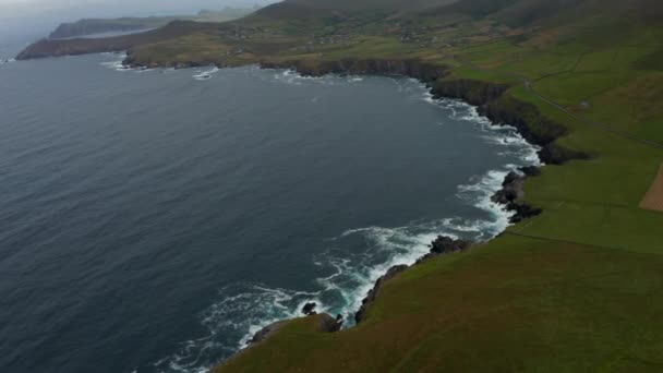 Luftaufnahme Küstenlandschaft. Wellen krachen auf die felsige Küste und bilden weißen Schaum. Grüne Weiden und Dörfer auf dem Land. Irland — Stockvideo