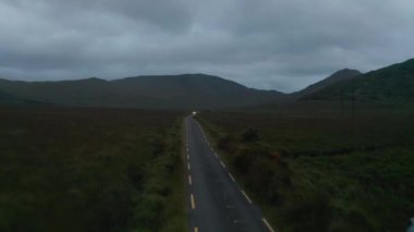 İleri, dar bir ülkenin üzerinde uçar ve arabalara çarpar. Alacakaranlıkta bulutlu bir gün. Arka planda dağ sırtı. İrlanda