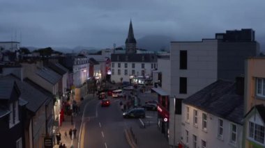 İleri uçaklar sokağın üstünde ve alacakaranlıkta kasaba meydanında uçar. Arabalar yolda gidiyor. Aydınlanmış ev cepheleri. Killarney, İrlanda