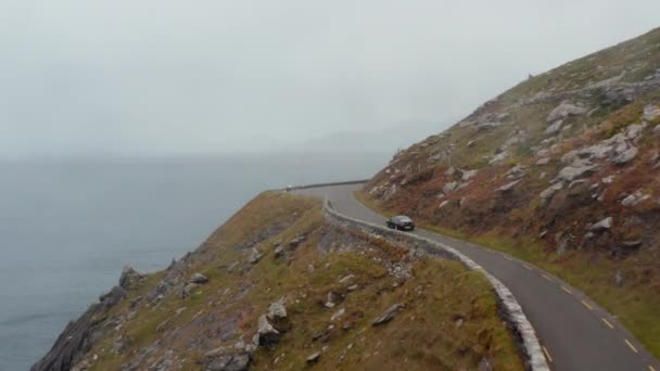 Přední sledování černého auta jedoucího po úzké, nechráněné pobřežní silnici vysoko na útesu. Mlhavý výhled na moře a kopce. Irsko — Stock video