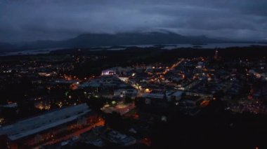 Akşam kasabasının üzerinden uç. Turuncu ışıklı caddelerde düşük trafik. Göl ve dağlar bulutlarla kaplı. Killarney, İrlanda