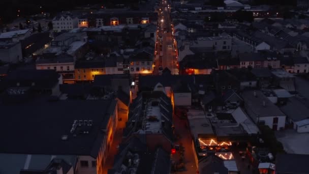 Forwards fly above illuminated street in town after sunset. Tilt up reveal of overcast sky and mountains in distance. Killarney, Ireland — Stock Video