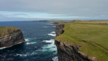İleri, yüksek ve dik uçurumların üzerinde uçar. Dalgalar kıyıya çarpıyor. Doğa manzarası soluyor. Kilkee Cliff Walk, İrlanda