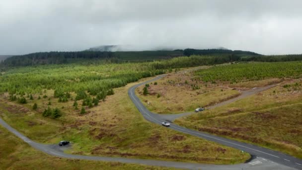 Nagranie z lotu ptaka z samochodu jadącego po drodze w górzystym leśnym krajobrazie. Wzgórza w chmurach w tle. Irlandia — Wideo stockowe