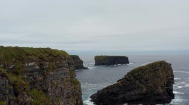 İleri doğru deniz kıyısı boyunca uçar. Yüksek kayalıklar, kayalık adalar ve dalgalar halinde adacıklar. Doğal manzarada hava görüntüsü. Kilkee Cliff Walk, İrlanda