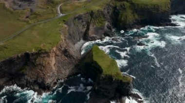 Deniz kıyısında yüksek açılı kaynar su manzarası. Dalgalar kıyıya çarpıp yuvarlanıyor. Yüksek dik yamaçların üzerindeki yeşil çayırlar. Kilkee Cliff Walk, İrlanda
