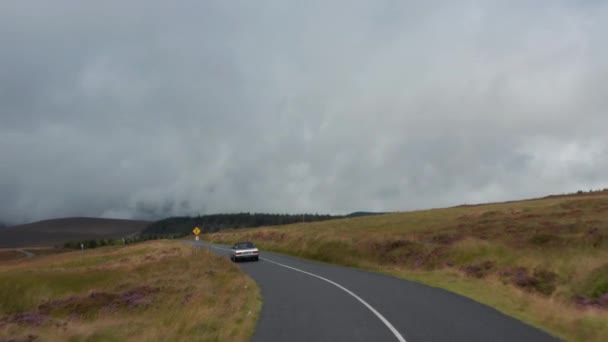 Przednie śledzenie zabytkowego samochodu jadącego po drodze otoczonego wrzosowiskami. Krajobraz górski ze wzgórzami pokrytymi chmurami. Irlandia — Wideo stockowe