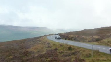 Dağlardaki bozkırlardan geçen yolda giden arabalar. Vadide alçak bulutlar olan yağmurlu bir hava. İrlanda