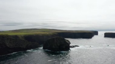 Deniz kıyısının ve dalgalı su yüzeyinin hava panoramik görüntüleri. Yukarıdaki yüksek dikey kaya duvarları ve otlaklar. Kilkee Cliff Walk, İrlanda