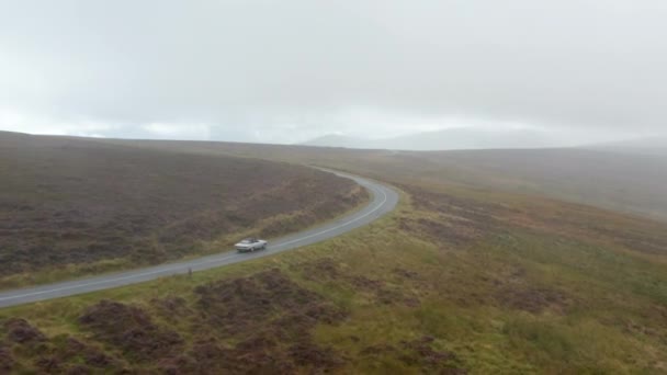 Coche deportivo vintage conduciendo por carretera mojada serpenteando entre pastizales en el campo. Día nublado en paisaje nebuloso. Irlanda — Vídeos de Stock