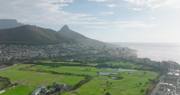 Volar por encima de césped verde exuberante en el campo de golf entre el desarrollo en la costa del mar. Montaña alta y plana en el fondo. Ciudad del Cabo, Sudáfrica — Vídeo de stock