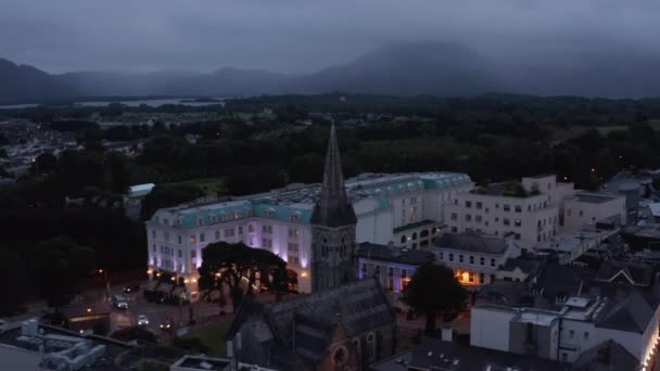 Historische stenen kerk met hoge toren en luxe hotelgebouw met verlichte gevel. Verkeer in avondstraten van de stad. Killarney, Ierland — Stockvideo