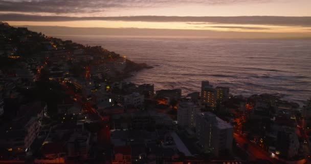 Fliegen Sie in der Abenddämmerung über Gebäude in Stadtvierteln an der Meeresküste. Wellenförmige Wasseroberfläche und bunter Himmel. Kapstadt, Südafrika — Stockvideo