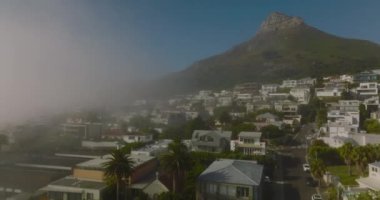 Turistik varış yerindeki evlerin üzerinden uç. İleride yüksek kayalık zirve var. Denizden sabah sisi yükseliyor. Cape Town, Güney Afrika