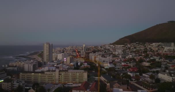夕暮れ時に街の上を飛ぶ。背景に通りや海に沿って複数階建てのアパートの建物。南アフリカ共和国ケープタウン — ストック動画