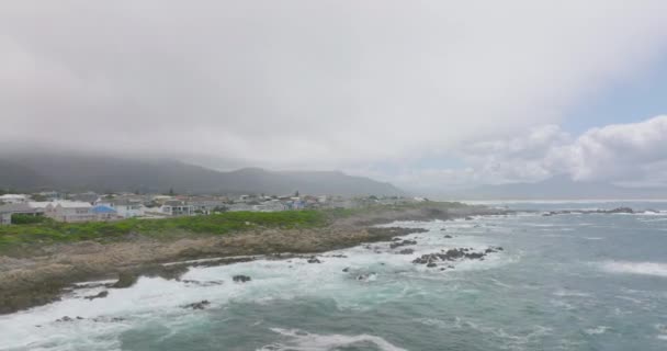Diapositive et panoramique des vagues qui s'écrasent sur les rochers de la côte. Maisons familiales en ville en arrière-plan. — Video