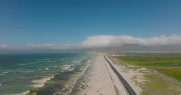 En arrière voler au-dessus de la côte. Route menant à travers le paysage sablonneux et les vagues roulant sur la plage. — Video