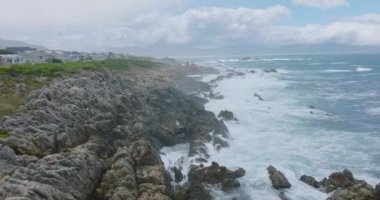 Kıyıdaki kayalara çarpan dalgaların üzerinden uç. Geriye doğru, yerleşim yerinin yakınlarındaki sert ve pürüzlü kayalık sahil kıyıları görünüyor..