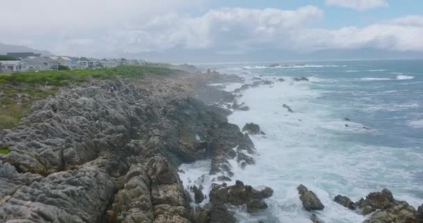 Vlieg boven golven die neerstorten op kustrotsen. Achterwaarts onthullen van ruige en grillige rotsachtige kust in de buurt van residentiële ontwikkeling. — Stockvideo