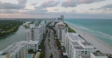 Alacakaranlıkta deniz kıyısındaki çok katlı binaların üzerinde uç. Apartman daireleri arasında çok şeritli yolda giden arabalar. Miami, ABD