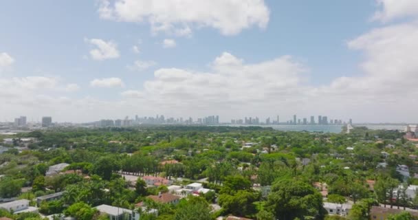 Forwards fly above residential neighbourhood with family houses and residences surrounded by greenery. Skyline with skyscrapers in distance. Miami, USA — ストック動画