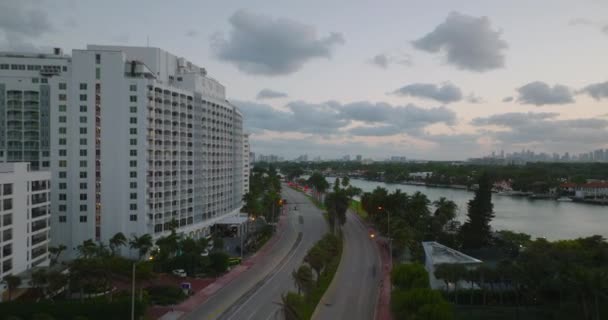 Передній політ над багатосмуговою дорогою веде через тропічне місто. Пальмові дерева, що махають вітром у сутінках. Повітряний вид на сучасне місто з багатоповерховими будинками. Маямі (США) — стокове відео