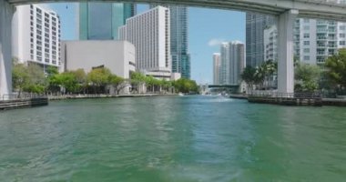 Modern şehirde köprü altından geçen motorlu tekne grubunu takip ediyoruz. Miami nehri ve şehir merkezindeki deniz kenarındaki binalar. Miami, ABD