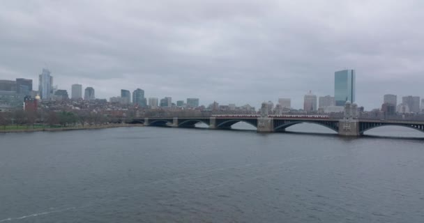 Red line train driving on Longfellow Bridge with urban borough in background. Forwards fly above rippled water surface. Boston, USA — Video
