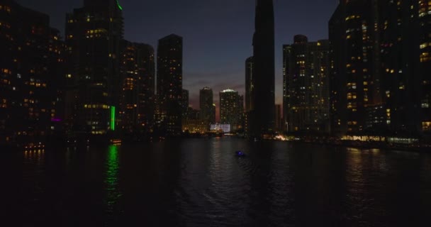 Low angle view of tall buildings with lighted windows around river. Forwards fly above water surface at night. Miami, USA — Stock Video