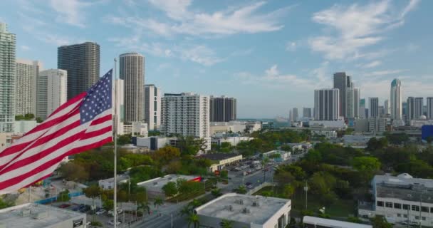 Vue aérienne de l'arrondissement urbain avec bâtiments et verdure. Le drapeau américain et les immeubles de grande hauteur en arrière-plan sont révélés. Miami, États-Unis — Video